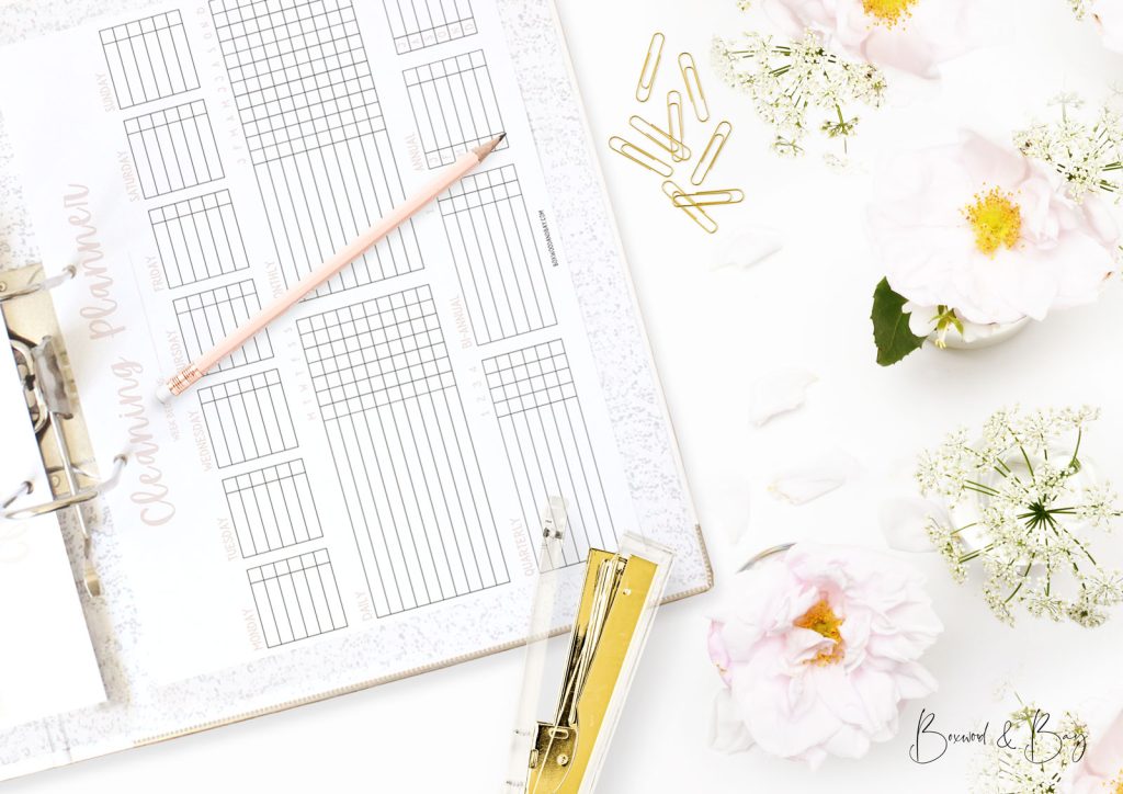 A top view image of an open lever arch binder on a desk surrounded by pretty pink and gold accessories including gold coloured paper clips, A gold and transparent stapler, a blush pink pencil and pretty pale pink and white flowers in small white china vases. Scattered petal litter the table top.