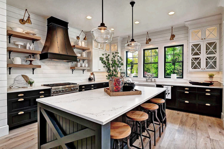 An image of a contemporary style farmhouse kitchen. Dark cabinets contrast with white marble worktops. Brass, copper and black metal highlights. Open natural wooden shelves, black window frames. Rustic style kitchen stools with black metal legs and wooden seats.