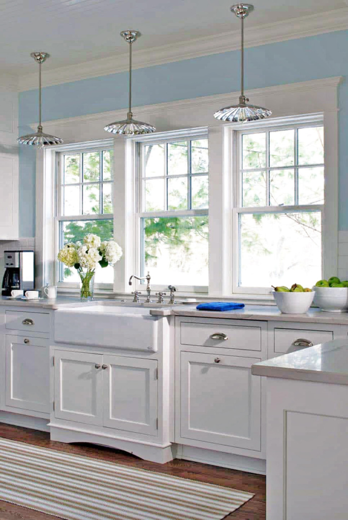 An image showing a country style farmhouse kitchen. Classic bespoke white cabinets, traditional white Belfast style sink. Pale blue walls and polished granite worktops complete the look.