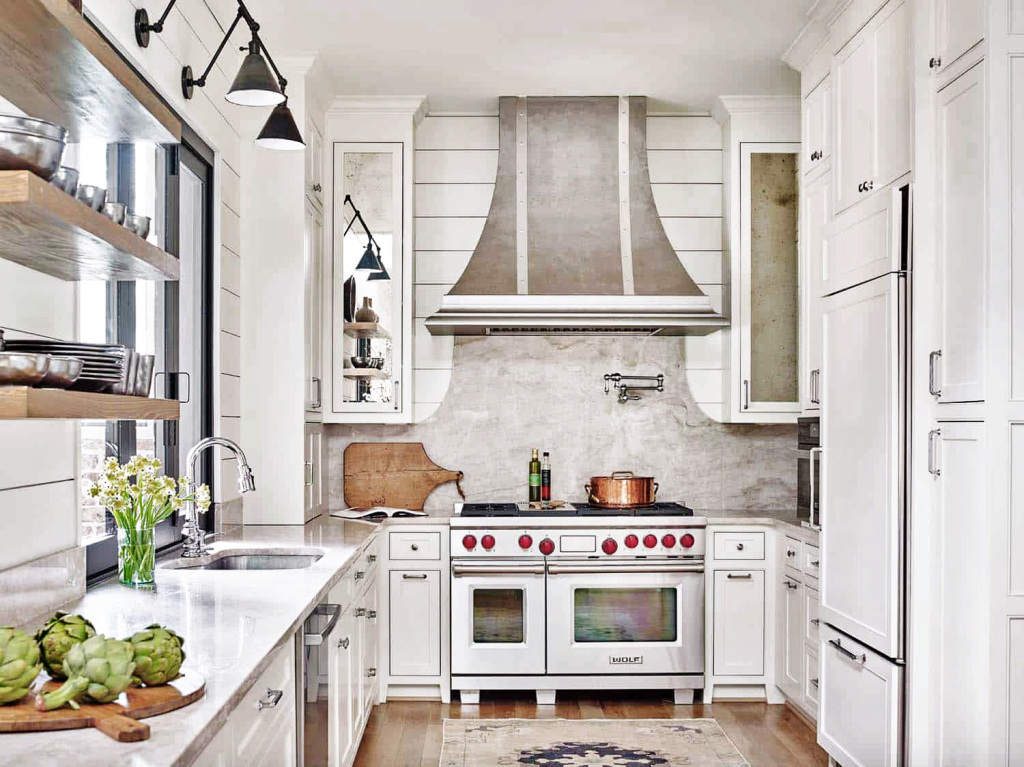 An image of a french inspired traditional country farmhouse kitchen with modern touches. A Wolf Range and its cooker hood steal the limelight. The cabinets are white and classic and simple. High level black angled wall lights provide interest.