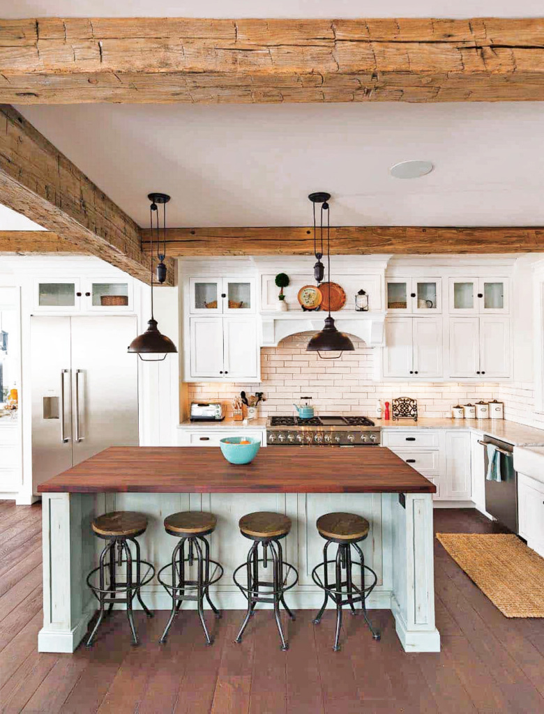 An image of a family designed open plan kitchen. Features large island for casual dining and meal prep. White cabinets and touches of mint green adds a highlight. Black industrial pendant light fittings hang above the island. Lots of exposed natural wooden beams and worktops complete the look.
