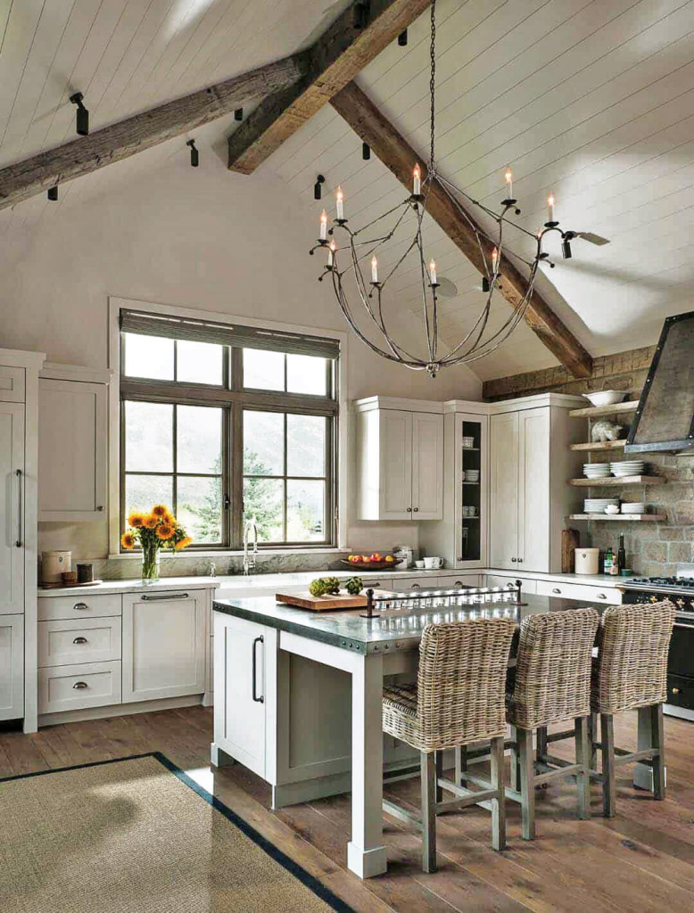 An image showing an open plan modern farmhouse kitchen with vaulted ceiling with exposed natrual timber beams. The cabinets are painted in antique ivory. An airy gunmetal electric candle effect chandelier gives the impression of age. Open chunky shelves and integrated appliances offer a sense of space.