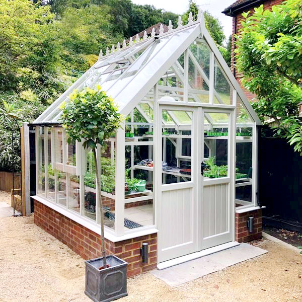 An image of a white greenhouse with a brick dwarf wall base, perfect for any flower garden.