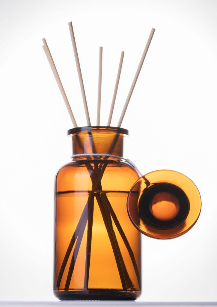 The photograph shows an amber diffuser bottle with it's lid attached at the neck by a piece of fine waxed twine. The lid is hanging down the side of the bottle. The bottle contains a scented liquid. There are six thin bamboo sticks placed in the bottle's liquid.