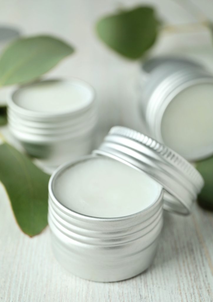 A photograph of a three small tins of handmade lip balm. The one in the foreground and in focus, has its screw top lid leaning against one side.