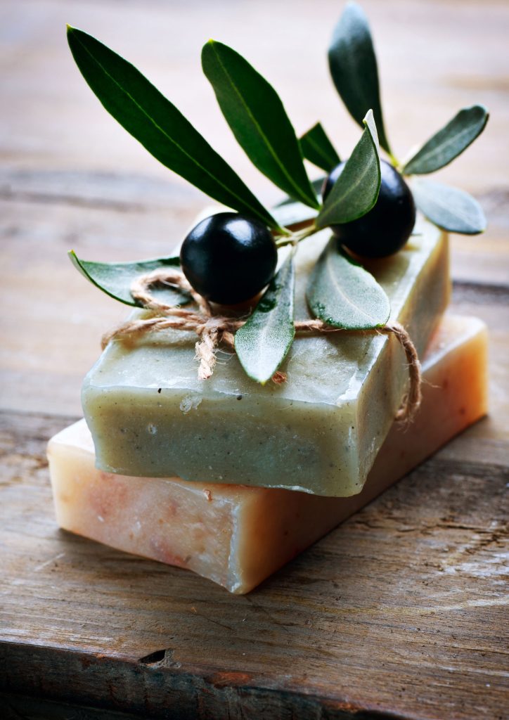 Photograph shows two bars of handmade olive soap (one on top of other), tied together with twine on a rustic wooden table. There is a small fresh olive sprig placed on top.