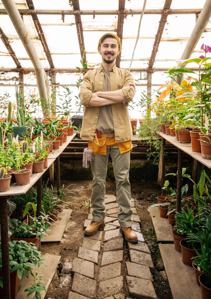 An image of a man standing in a vintage greenhouse surrounded by potted plants.