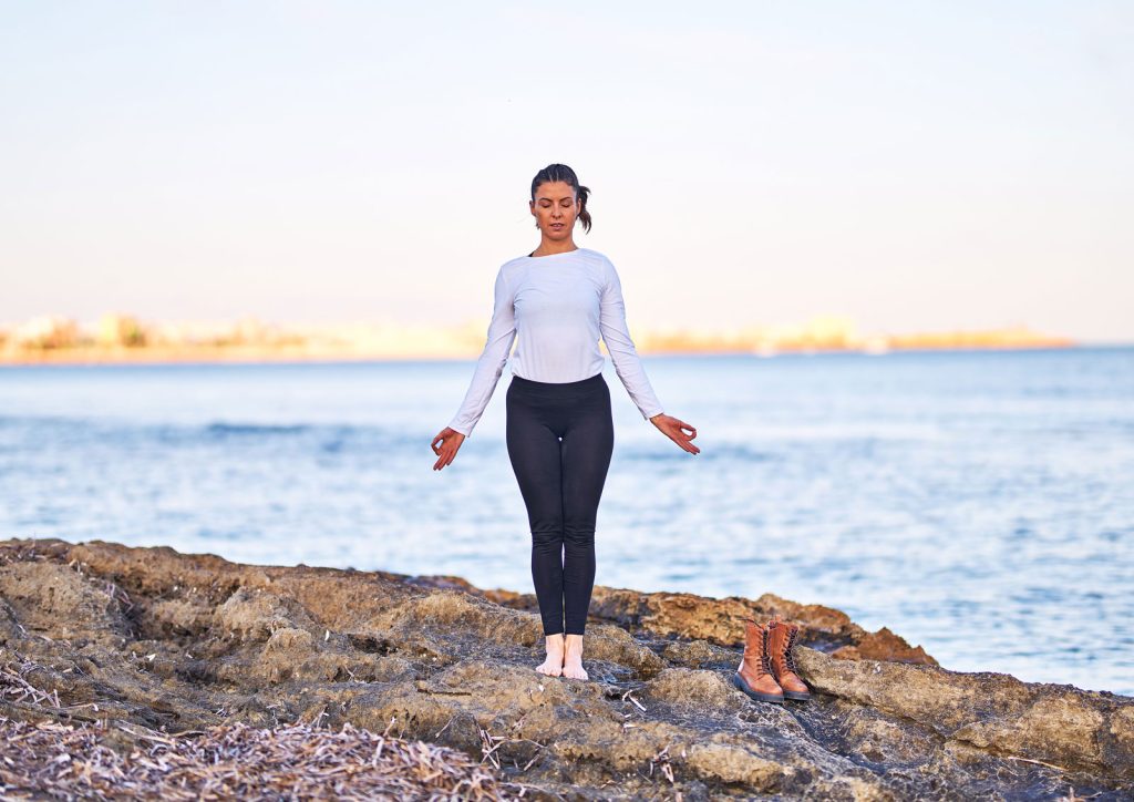 Photograph of a woman presenting yoga's Mountain Pose on a rocky outcrop against an ocean backdrop.