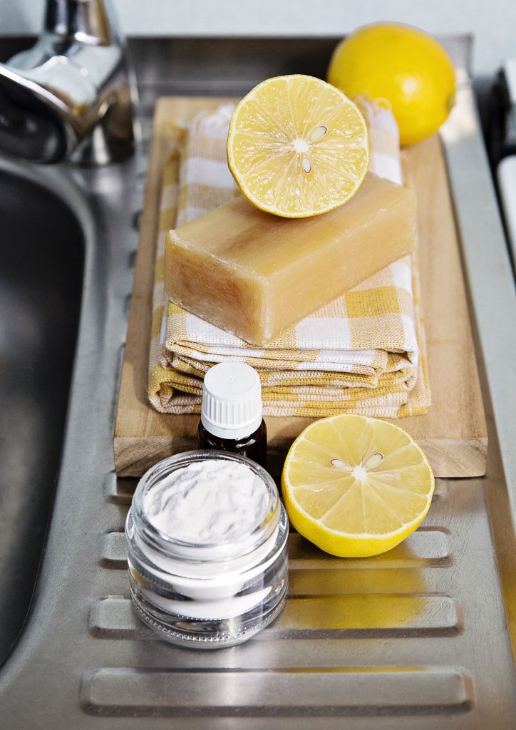 A photograph depicting natural cleaning products: lemons, bicarbonate of soda, essential oil and a bar of castile soap.