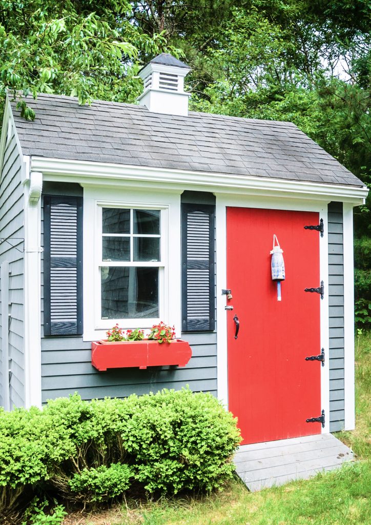 A photograph of a cute flower garden shed painted in a blue-grey shade, with a bright red door and matching window box placed under the front window.