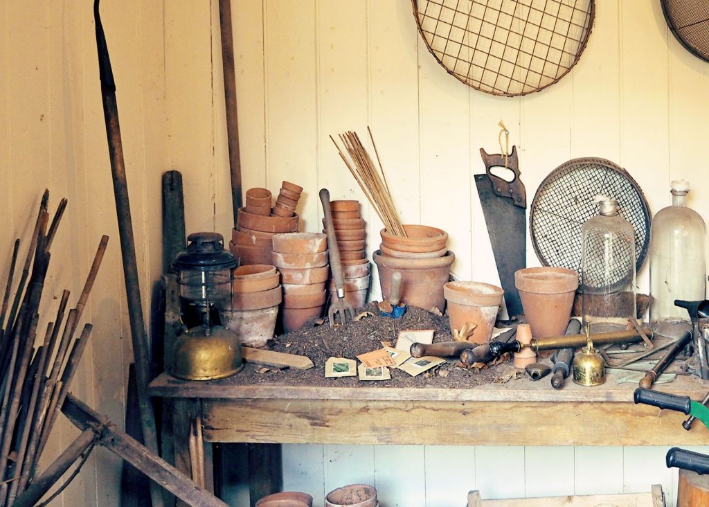 The image shows the interior of a rustic potting shed. There are flower garden implements strewn about and loose seeds on the potting bench, which is covered in spilt soil.