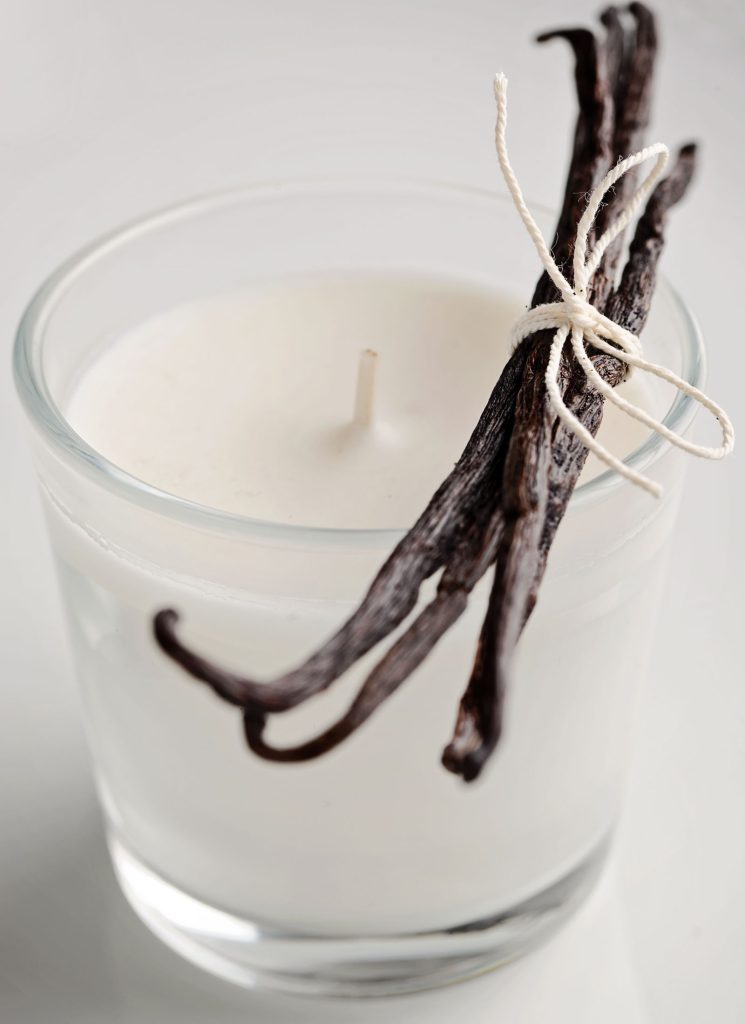 The photograph depicts a glass votive candle with white wax. There is a bundle of vanilla pods tied together with string, place across the top.