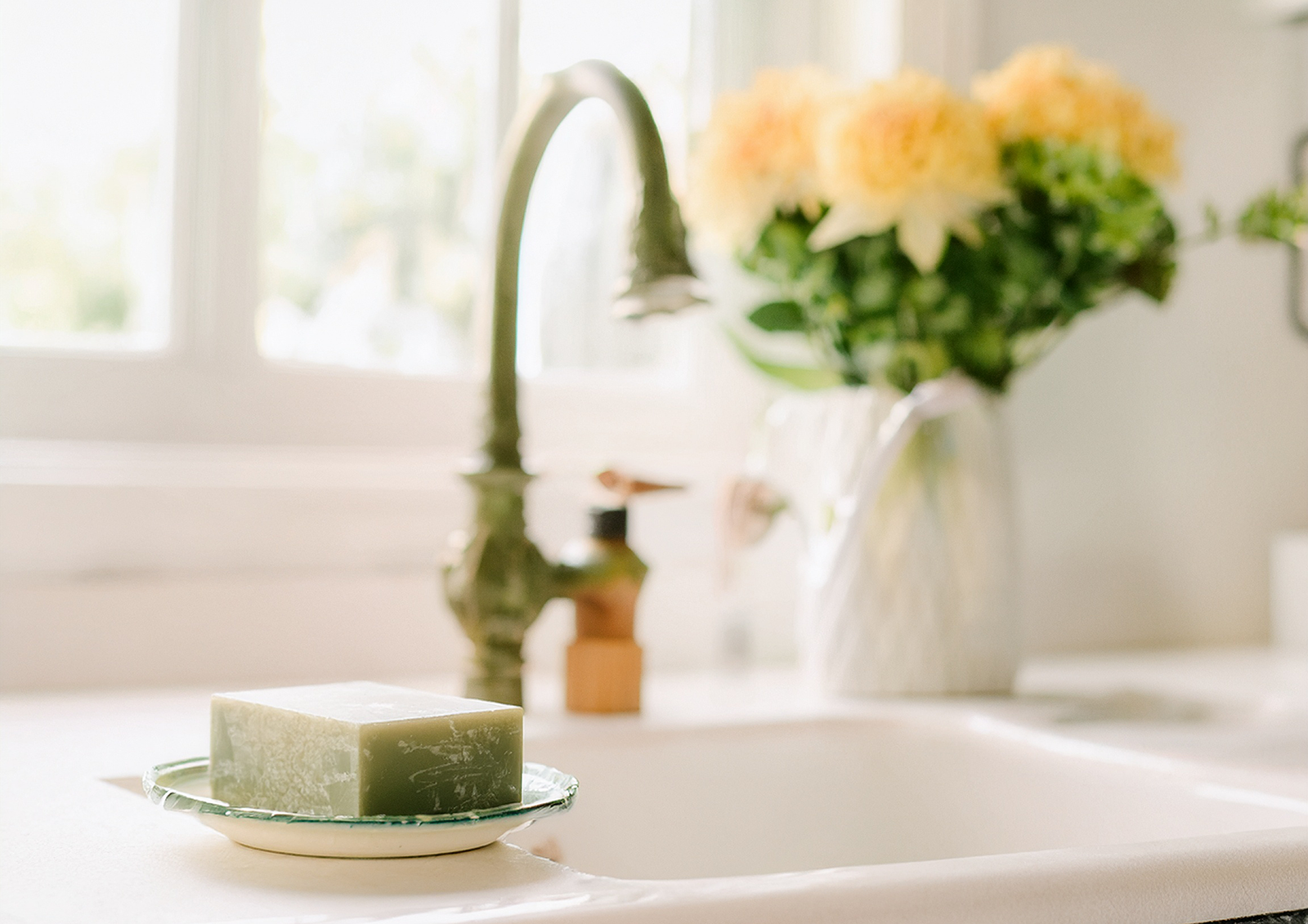 Image of a white sink featuring a small porcelain dish with a block of olive soap in the foreground. Blurred in the background is a vase of peach coloured flowers, a verdi-gris tap and an amber bottle of hand cream.