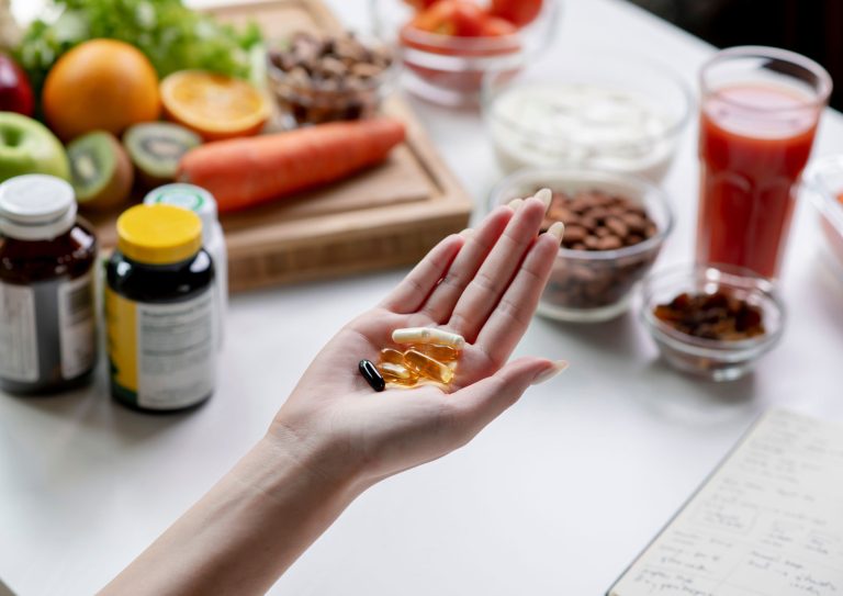 Photo of a woman's open hand, containing a small pile of supplement capsules. Fresh fruit, vegetables, juice, nuts and vitamin supplements for immune system containers can be seen in a blurred background.