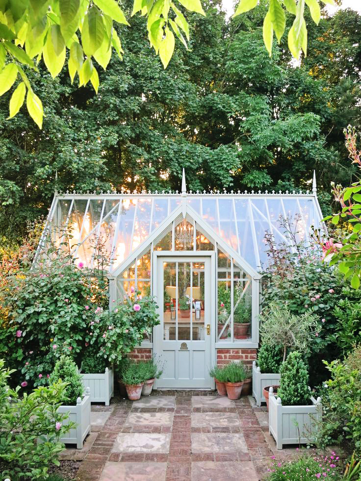 An image of a pretty Victorian orangery-style greenhouse painted in a shade of blue-grey. There is a feature path of paving stones and pea gravel, flanked by handsome planters, leading to the greenhouse, perfect for a flower garden.