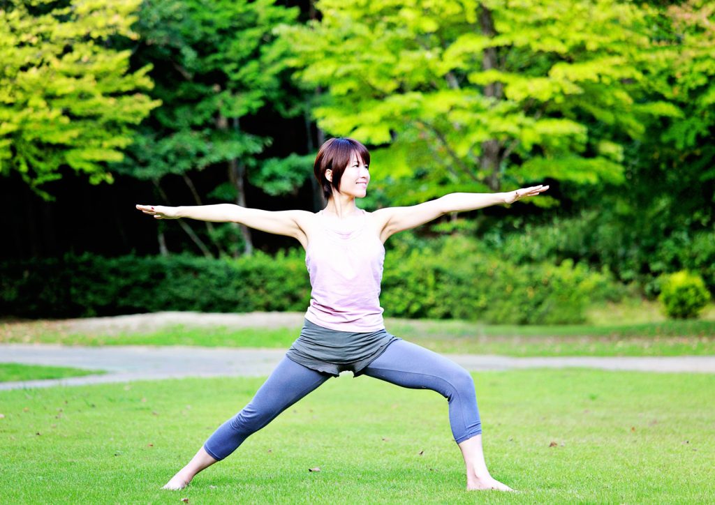 Photograph of a woman presenting yoga's Warrior II pose in a parkland setting against a backdrop of lush trees.