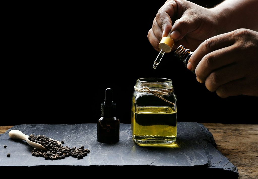 Photograph shows a person (out of sight) putting drops of essential oil from a pipette into a glass container of a carrier oil.