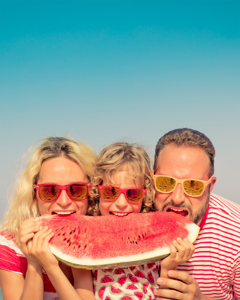 An image of a woman, a child and a man standing next together, are eating a large wedge of watermelon with the ocean as a backdrop.