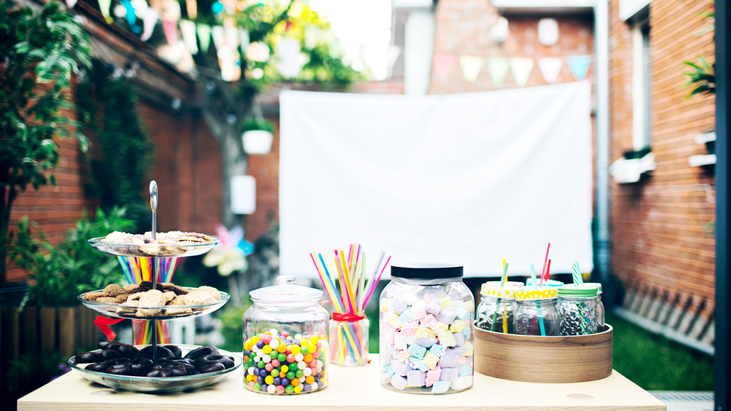 An image of an outdoor garden movie night set-up. There is a hung white sheet in the background and in the foreground is a table with an assortment of sweets and snacks.