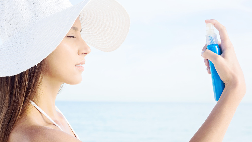An image of a woman at the beach wearing a white wide brim sun hat raising a spray container to spray face.