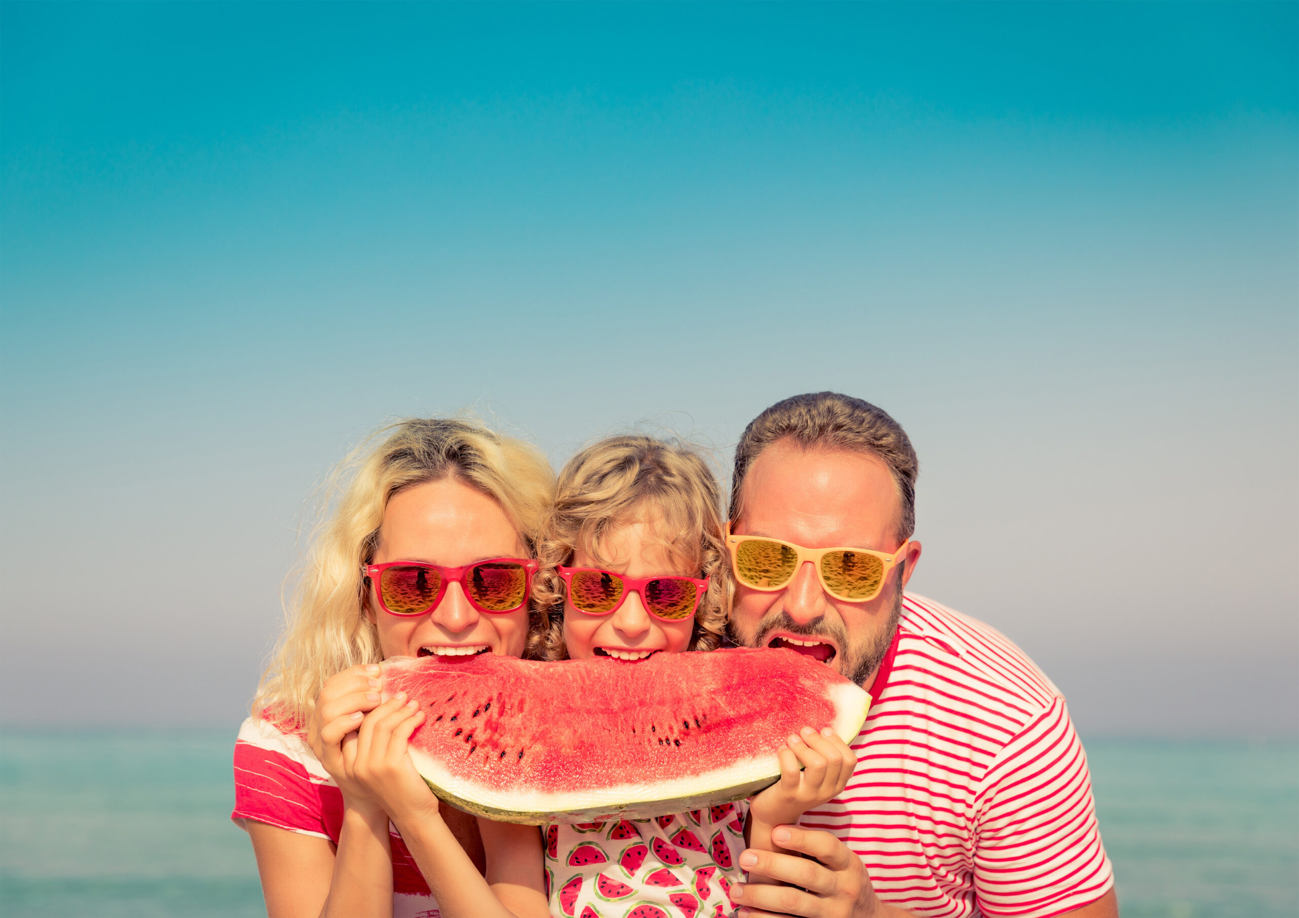 An image of a woman, a child and a man standing next together, are eating a large wedge of watermelon with the ocean as a backdrop. Summertime.