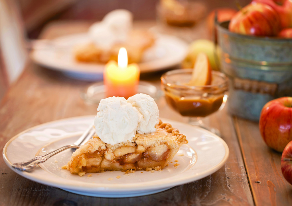 Image showing a slice of homemade apple pie topped with two scoops of vanilla ice cream. The setting is rustic and warm with a mellow candle burning, small metal teal bucket containing red apples, a small glass bowl of soft caramel for dipping apple wedges.