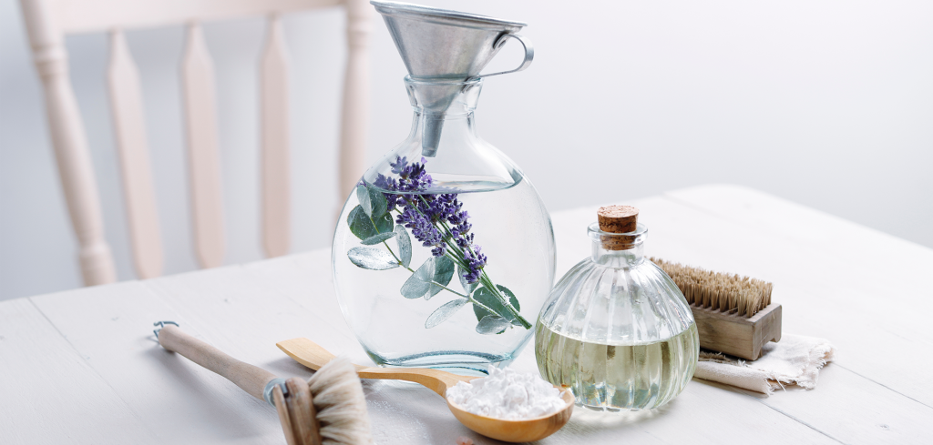 An image of a grouping of natural ingredients to make natural cleaning products siting on a rustic bleached table. There is a oval shaped glass jug containing white vinegar with sprigs of eucalyptus and lavender. There is a small rustic galvanised funnel sitting in the neck of the jug. Also on the table are: a small rounded ribbed jar with cork bung in the neck, containing essential oil blend; a wooden spoon with bicarbonate of soda; pink Himalayan salt crystals are scattered on the table top; There is a wooden and natural dish washing brush and a wooden and natural bristle nailbrush sitting on a small folded piece of natural cotton.
