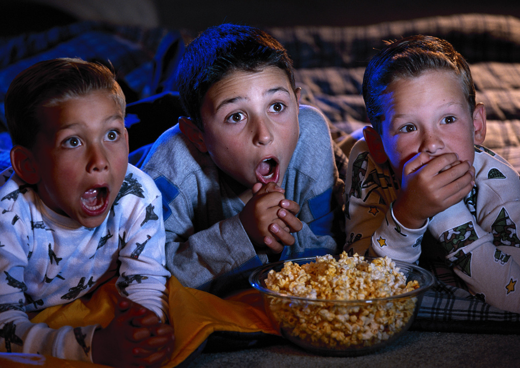 An image of three boys wearing pjs laid on the floor in front of a glass bowl of popcorn watching a movie with astonished expressions on their faces.